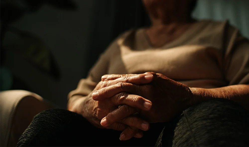 Close-up of a seated person with hands clasped together in their lap, softly lit in a dim interior setting, with focus on the hands and blurred upper body in the background.
