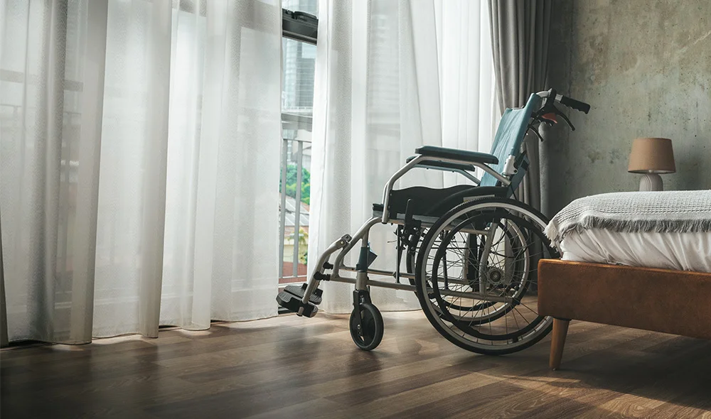 A wheelchair positioned beside a neatly made bed in a softly lit bedroom, with sheer curtains filtering daylight through large windows and a small bedside lamp on a wooden nightstand.