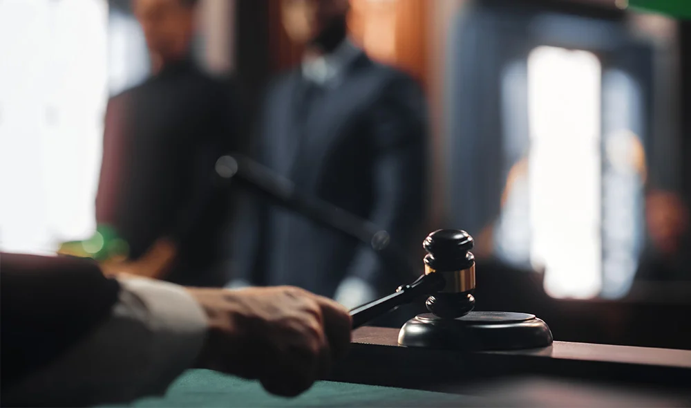 Close-up of a judge’s hand holding a wooden gavel poised above a sound block in a courtroom setting. In the softly blurred background, several people in formal attire stand near a witness area, with warm lighting and a green desk lamp illuminating the sce