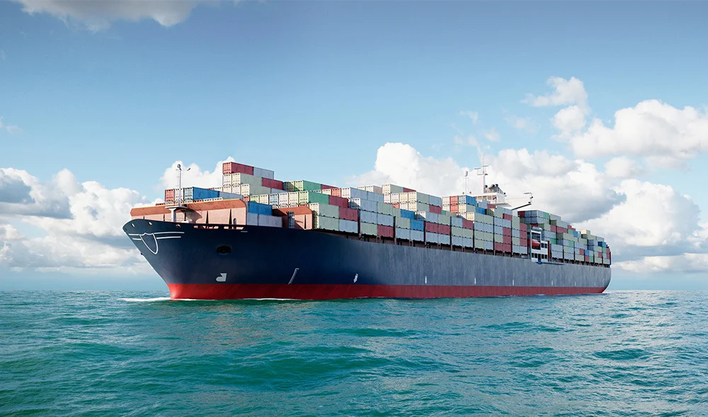 Large cargo ship traveling across open water, loaded with multicolored shipping containers stacked high on its deck, under a bright blue sky with scattered clouds.