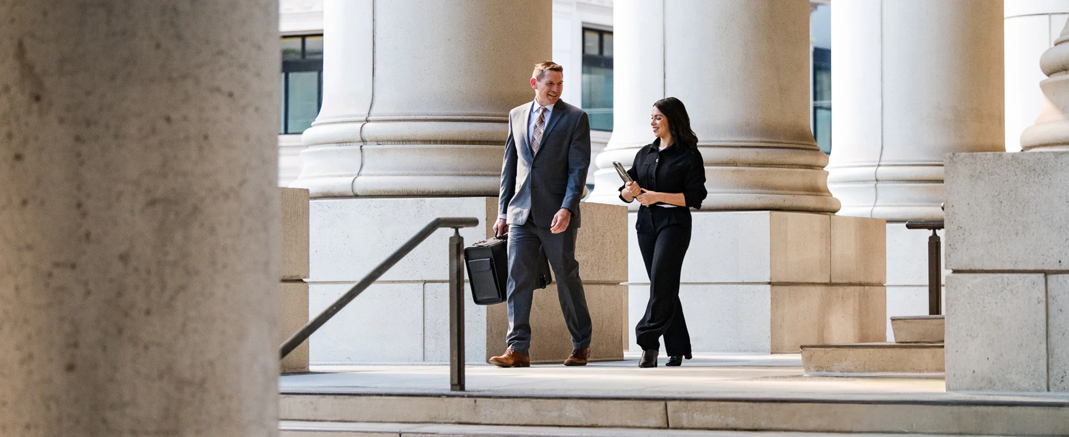 Two business professionals walk side by side through a colonnaded civic or office building. One carries a briefcase while the other holds a folder, with large stone columns, steps, and railings framing the bright outdoor walkway.