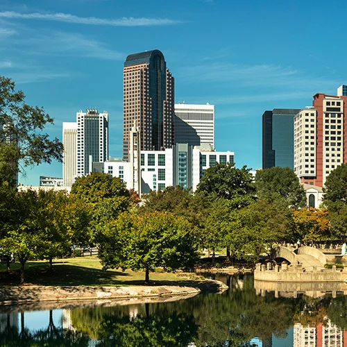 Charlotte skyline viewed from Marshall Park, featuring modern office buildings reflected in a calm pond, framed by green trees and a clear blue sky.