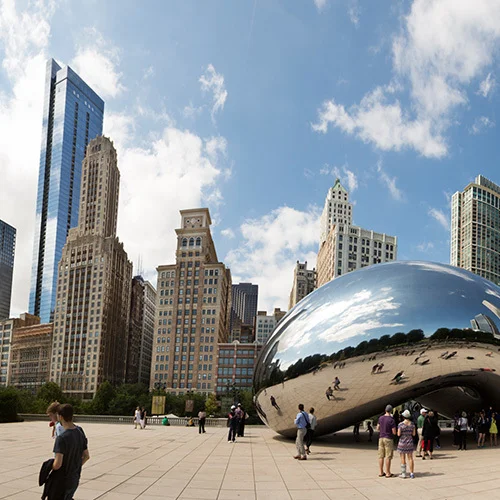 Chicago skyline with Cloud Gate sculpture (the Bean) in Millennium Park, surrounded by visitors, tall skyscrapers, trees, and a bright blue sky with scattered clouds.
