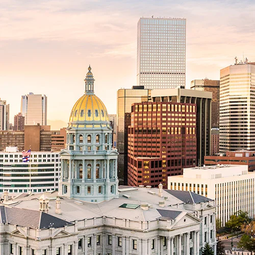 Downtown Denver skyline with the Colorado State Capitol’s gold dome in the foreground, surrounded by modern office towers under a soft, pastel sunset sky.