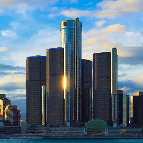 Detroit skyline at sunset featuring the Renaissance Center’s cylindrical glass towers along the Detroit River, with dramatic clouds and warm light peaking through the buildings.