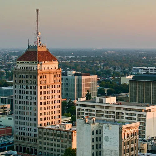 Fresno city skyline at dusk highlighting a high-rise building with a red-tiled roof and rooftop antennas, surrounded by low-rise offices and trees stretching into the distance.