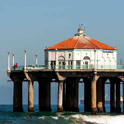 Hermosa Beach pier extending over the Pacific Ocean with a round pavilion topped by a red tile roof, ocean waves breaking below, and a clear blue sky in the background.