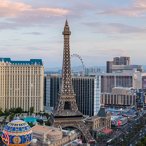 Panoramic view of the Las Vegas Strip at sunset, featuring the Paris Las Vegas Eiffel Tower replica, the Bellagio fountains and lake, and surrounding resort hotels under a soft, glowing sky.