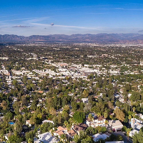 Aerial panoramic view of Woodland Hills, California, showing a sprawling residential neighborhood with tree-covered streets and homes stretching toward distant mountains beneath a clear blue sky.
