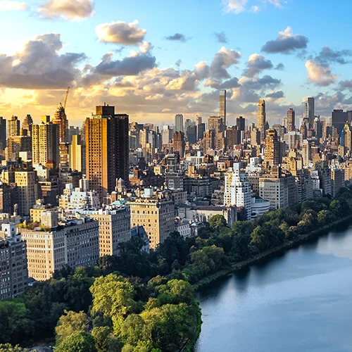 “Panoramic view of Manhattan skyline at sunset, with Central Park’s tree-lined reservoir in the foreground and tall skyscrapers rising behind under a partly cloudy sky.
