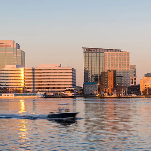 Waterfront skyline of downtown Norfolk at golden hour, showing modern office buildings and hotels reflected in calm harbor water with a small boat passing in the foreground.