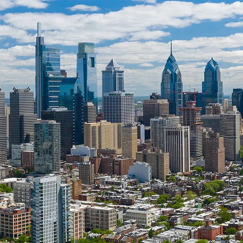 Panoramic aerial view of downtown Philadelphia, showing dense city blocks, modern skyscrapers including glass towers, and the Schuylkill River with bridges curving through the foreground under a partly cloudy blue sky.