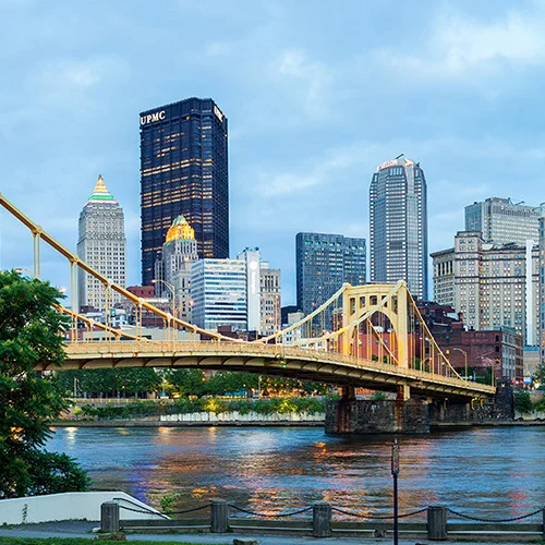 Wide view of downtown Pittsburgh at dusk, with yellow suspension bridges crossing a river in the foreground and a lit skyline of high-rise buildings reflected in the water beneath an overcast evening sky.