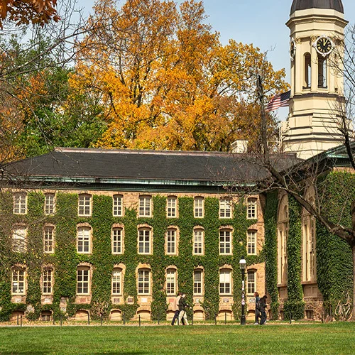 Historic stone academic building at Princeton University covered in ivy, with a clock tower and arched windows, surrounded by autumn trees with yellow and orange leaves and a grassy lawn in the foreground.