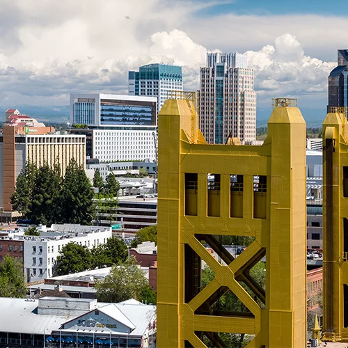 Downtown Sacramento skyline with the golden towers of the Tower Bridge in the foreground, surrounded by modern office buildings and tree-lined streets under a bright sky with large white clouds.