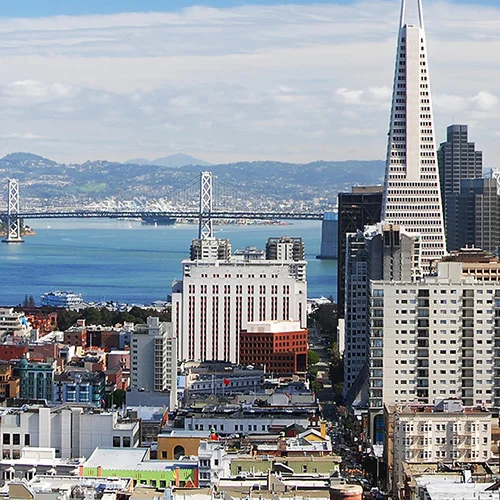 Wide view of downtown San Francisco showing the Transamerica Pyramid among dense high-rise buildings, with the San Francisco–Oakland Bay Bridge and blue bay water visible in the background.