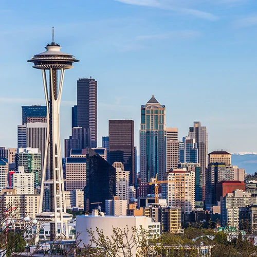 Panoramic view of Seattle featuring the Space Needle rising above downtown buildings, with Mount Rainier visible in the distance and Elliott Bay and port cranes along the waterfront under a clear blue sky