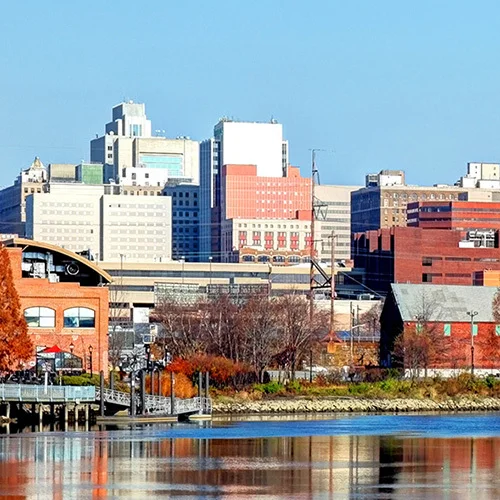 Panoramic view of Wilmington, Delaware’s riverfront skyline, showing modern office buildings and brick structures reflected in calm water, with autumn-colored trees and tall grasses along the shoreline beneath a clear blue sky.