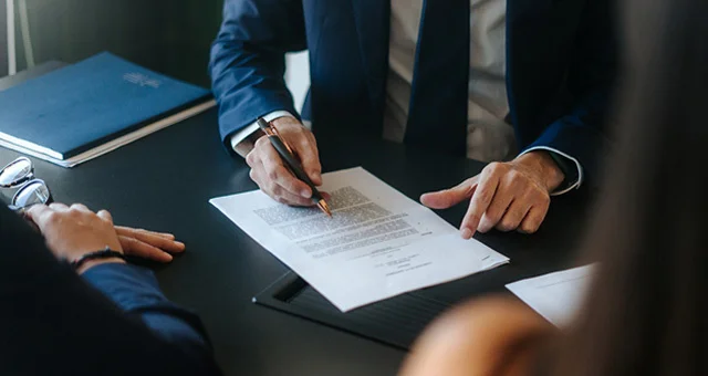 A close-up view of a professional workspace shows two individuals seated across a desk reviewing documents together. One individual’s hand holds a pen and points to a sheet of paper on a clipboard, while another individual’s hand gestures to it.