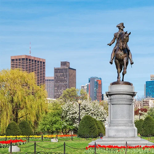 Paul Revere statue in a Boston Public Garden framed by blooming trees, with mid- and high-rise city buildings under a clear blue sky.