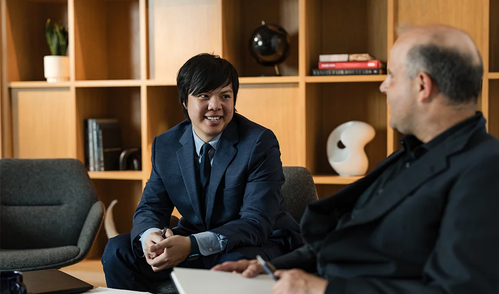 Two people sit across from each other at a desk in a professional office setting. One person, wearing a dark outfit, leans forward slightly while holding a pen and reviewing documents on the table. The other person is seated opposite.