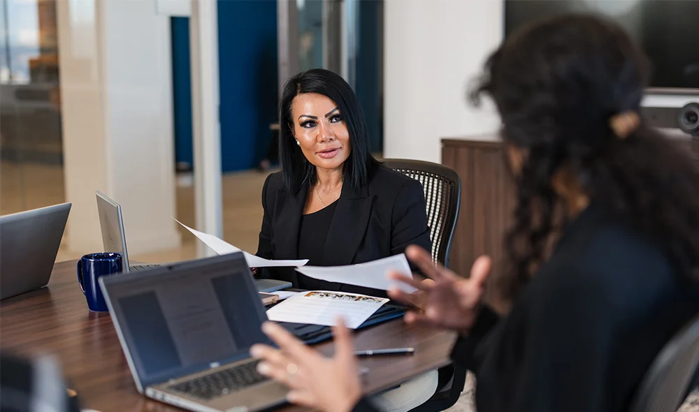 Two people sit across from each other at a desk in a modern office, engaged in a professional conversation. One person sits behind an open laptop while extending a hand across the table, and the other person reaches forward to meet the gesture.