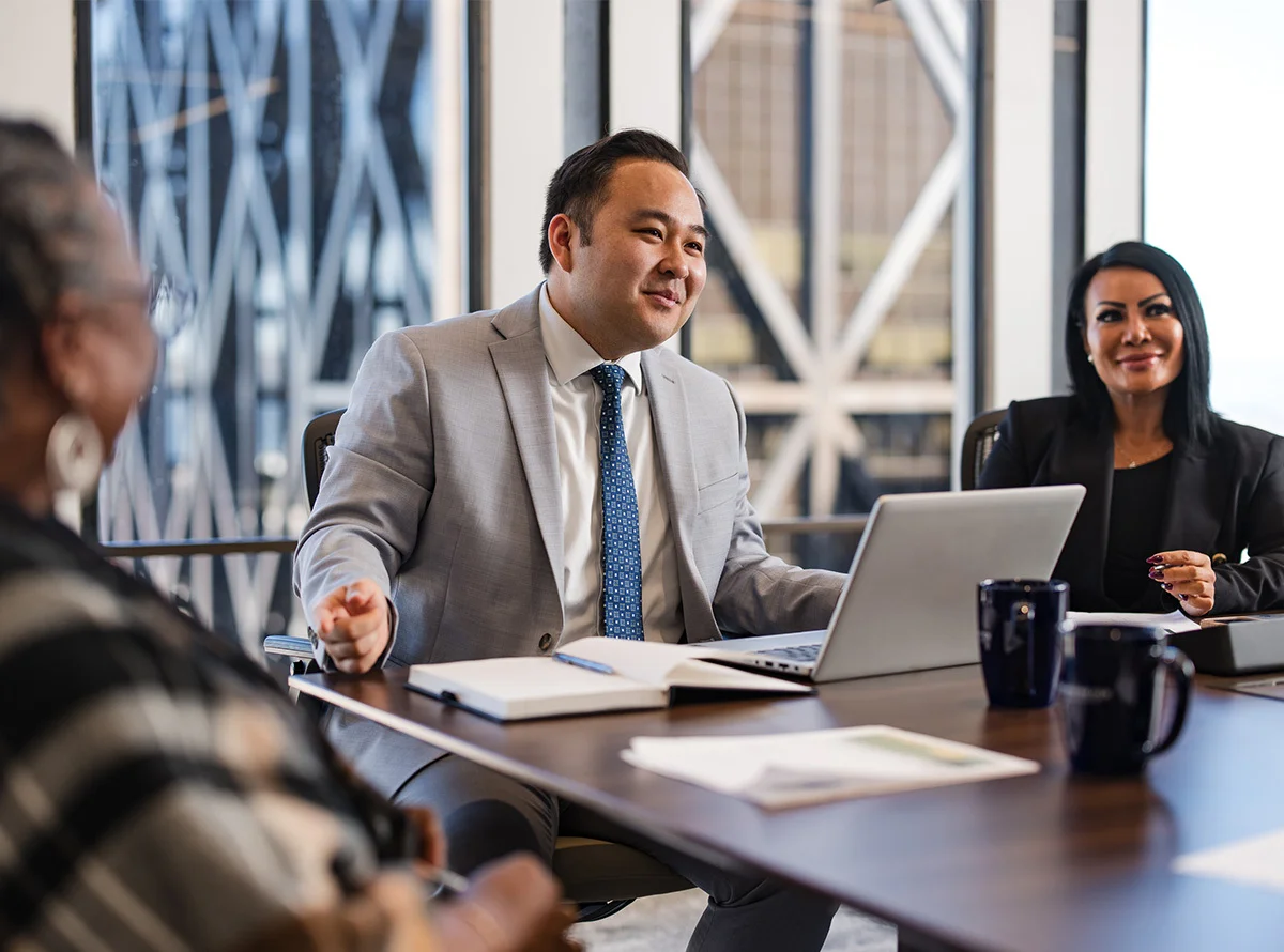 Three business professionals sit at a conference table in business professional attire, with coffee mugs on the table and a notebook opened, with natural lighting in large glass windows.