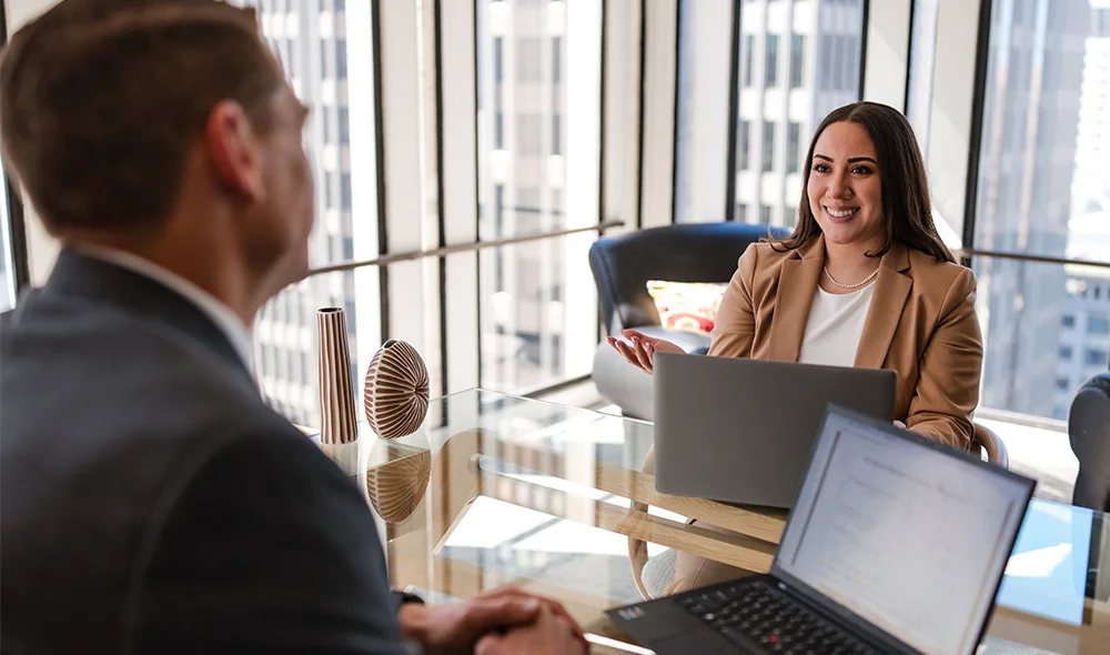 A professional meeting takes place in a bright office with large windows showing a city skyline. Two people sit across from each other at a table with open laptops and documents. One person gestures while speaking, with a coffee mug and papers nearby.