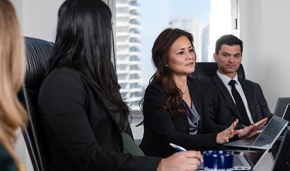 Three individuals are seated side by side at a desk in a modern office environment, participating in a professional meeting or interview. One person sits in the foreground with their back partially facing the camera, with two individuals across the table.