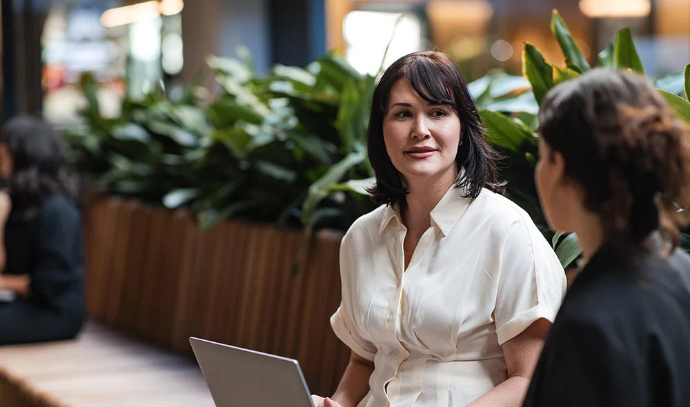 A person in a white blouse stands in a warmly lit, plant-filled indoor space, engaged in conversation with others. A laptop sits open on a nearby table alongside takeaway coffee cups and a notebook. Shelving and decorative items are in the background.