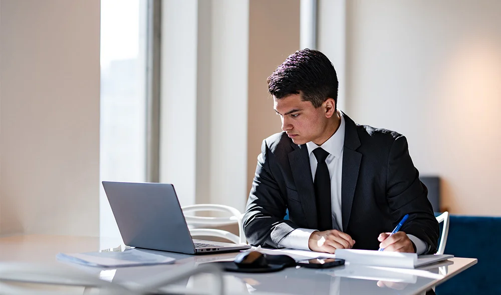 A person in formal business attire sits alone at a desk in a minimalist office setting, working on a laptop. Papers and documents are spread across the desk alongside a notebook and pen. The individual looks downward toward the laptop or documents.