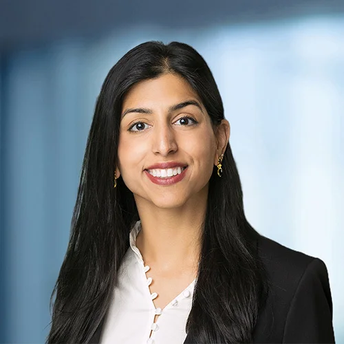 Professional headshot of Iman Eikram with long dark hair, wearing a black blazer and a light-colored blouse with a subtle scalloped detail at the neckline. The subject is facing forward against a softly blurred blue and gray background.