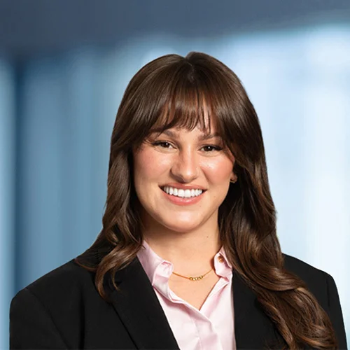 Professional headshot of Emmy Hoskins with shoulder-length brown hair and bangs, facing the camera against a softly blurred office-style background. Emmy is wearing a black blazer over a light pink collared shirt and a thin gold necklace. The lighting is