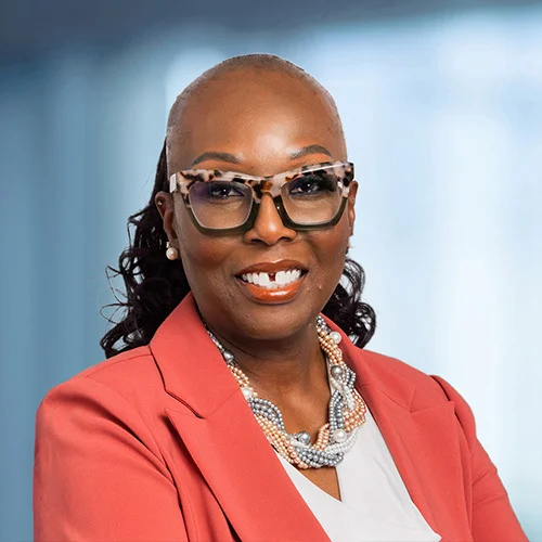 A professional headshot of Tina Samuel-Priestly wearing a coral blazer, light-colored top, and layered pearl necklace. The individual has short, closely styled hair, wears large patterned eyeglasses, and stands against a softly blurred blue-gray backgroun