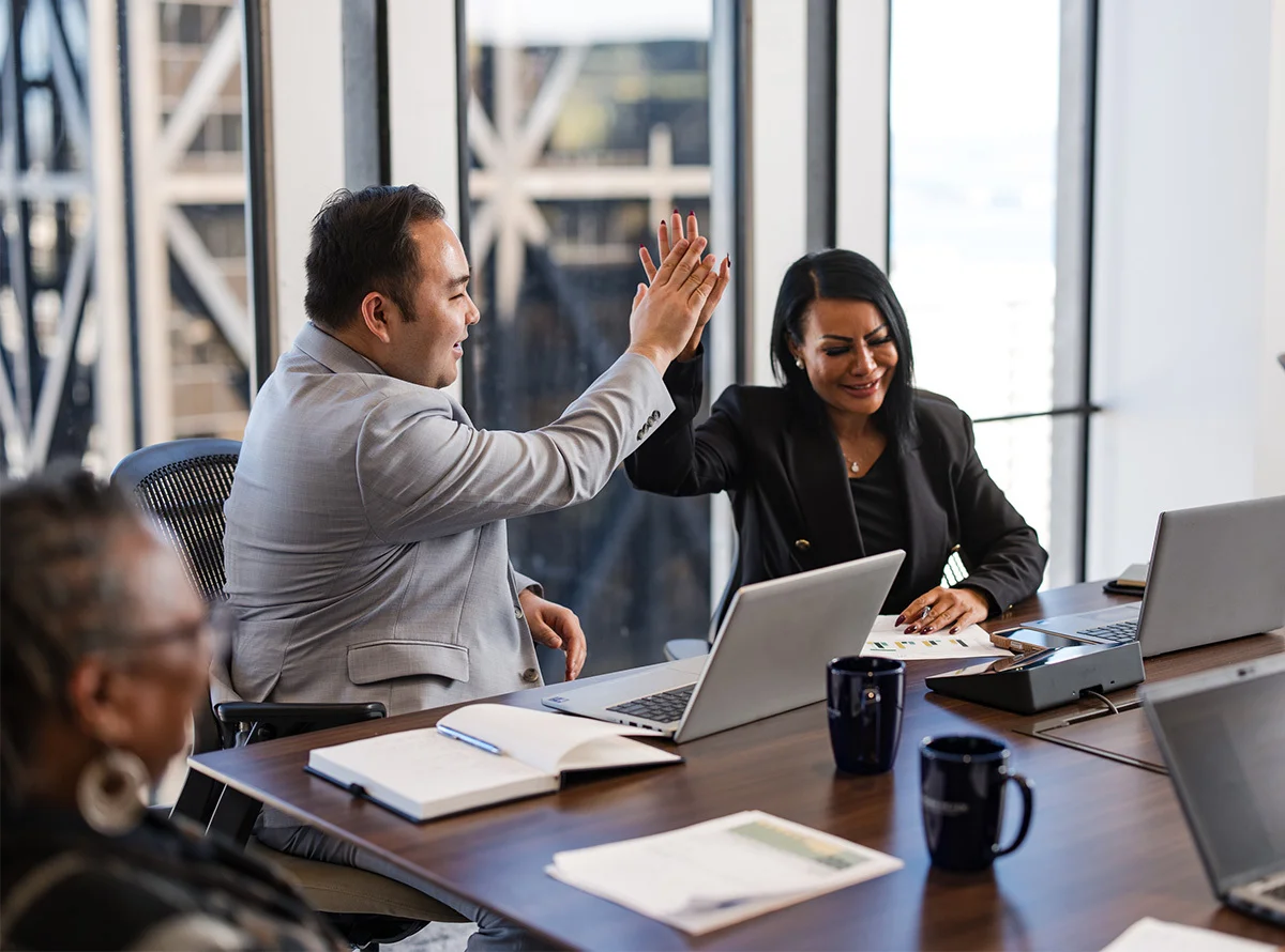 Two coworkers seated at a conference table exchange a high-five over open laptops. Papers, notebooks, and coffee mugs are arranged on the table. Large windows reveal a cityscape outside, and another person is partially visible in the foreground observing