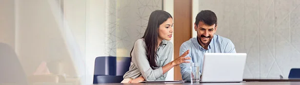 Two professionals seated at a conference table review information together on a laptop, collaborating in a modern office setting. A glass of water and notepad sit on the table beside them.