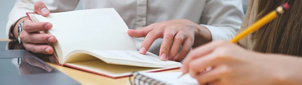 Close-up of two people working together, with one person pointing to an open notebook while the other takes notes with a pencil, illustrating collaboration and mentorship.