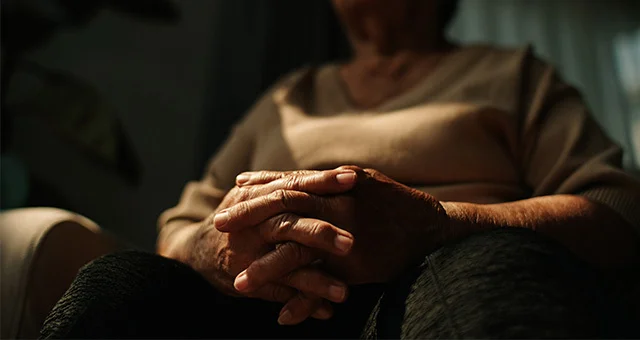 Close-up of a seated person with hands clasped together in their lap, softly lit in a dim interior setting, with focus on the hands and blurred upper body in the background.