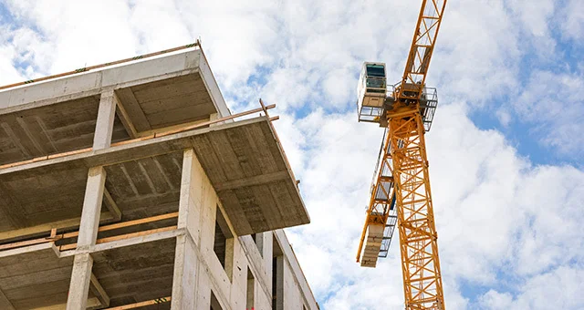 Wide-angle view of a construction site featuring a partially built concrete structure with multiple open floors and support columns. A tall yellow tower crane stands beside the building, extending into a partly cloudy sky, highlighting active development