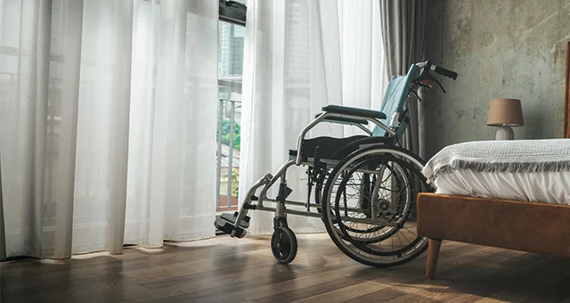 A wheelchair positioned beside a neatly made bed in a softly lit bedroom, with sheer curtains filtering daylight through large windows and a small bedside lamp on a wooden nightstand.