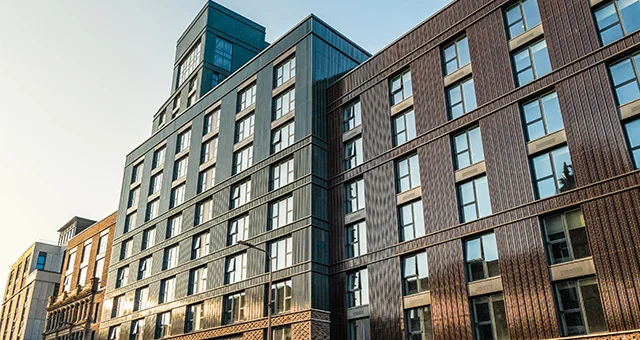 Modern mid-rise apartment or condominium building with a mix of brick and glass exterior, featuring large windows and varied facade textures. The structure rises several stories high under a clear sky, with neighboring buildings partially visible along th
