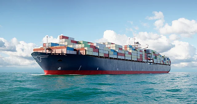 Large cargo ship traveling across open water, loaded with multicolored shipping containers stacked high on its deck, under a bright blue sky with scattered clouds.