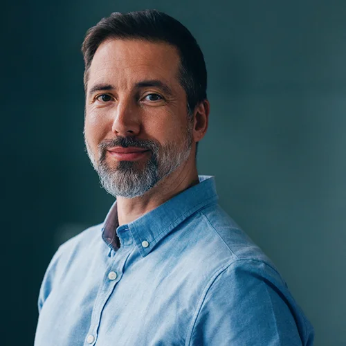 Professional headshot of a person wearing a light blue button‑down shirt, photographed from the chest up against a dark, softly lit background.