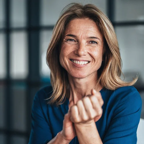 Portrait‑style image of a person wearing a blue top, seated with hands clasped in the foreground, photographed indoors against a softly blurred background.