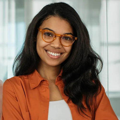 Portrait‑style image of a person wearing an orange button‑down shirt over a light top, photographed from the chest up indoors against a softly lit background.