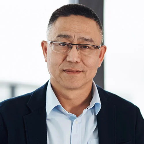 Portrait‑style image of a person wearing a dark blazer over a light button‑down shirt, photographed from the chest up indoors against a bright, softly blurred background.