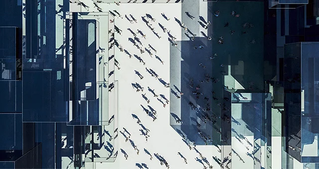 Overhead view of a modern urban plaza with geometric architectural structures casting shadows, where numerous people walk across an open space, creating patterns of movement and contrast between light and shade.