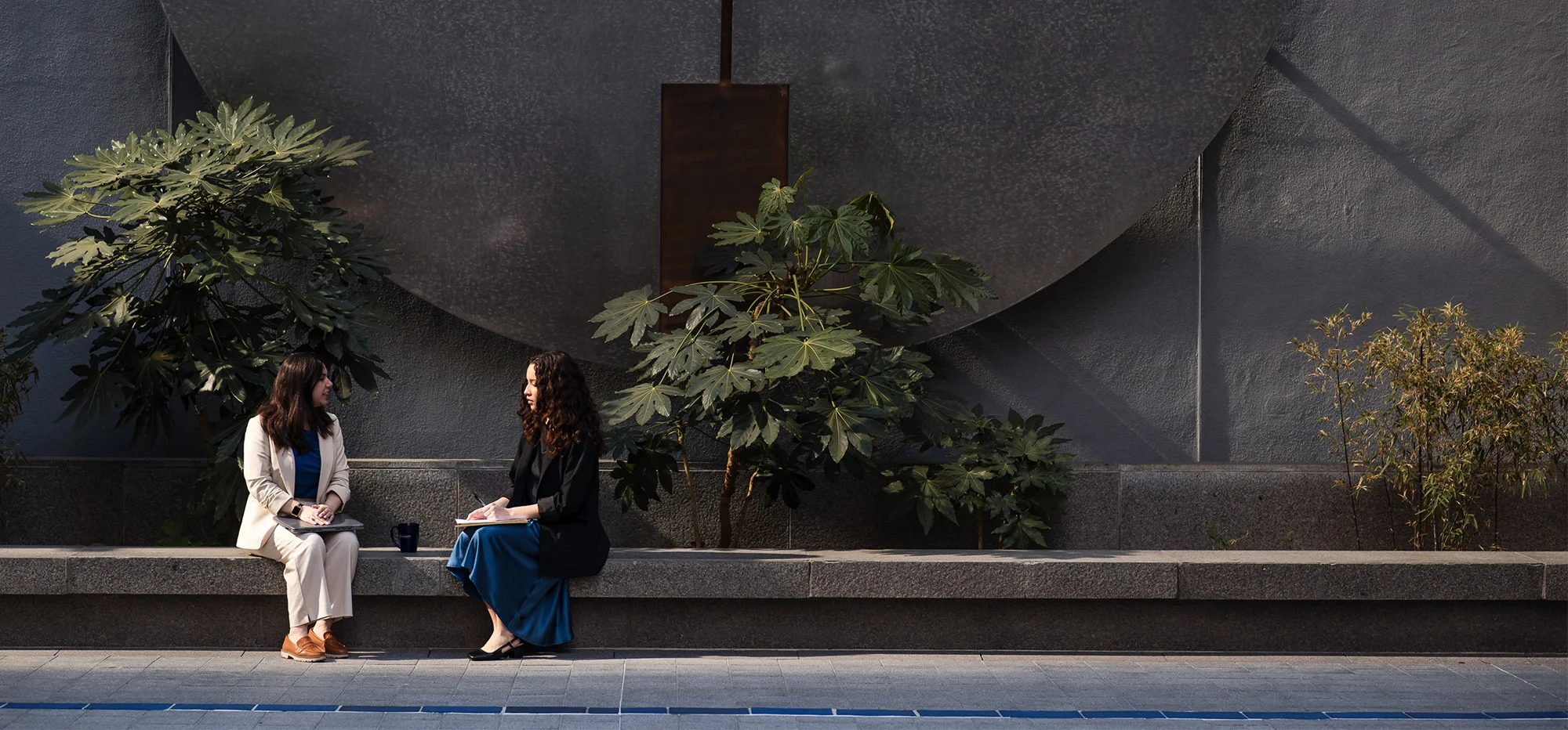 Two professionals sit on a long stone bench in an outdoor courtyard, meeting and reviewing notes beside a laptop and coffee mug. Large leafy plants and a textured architectural wall create a calm, modern setting, with natural sunlight casting soft shadows