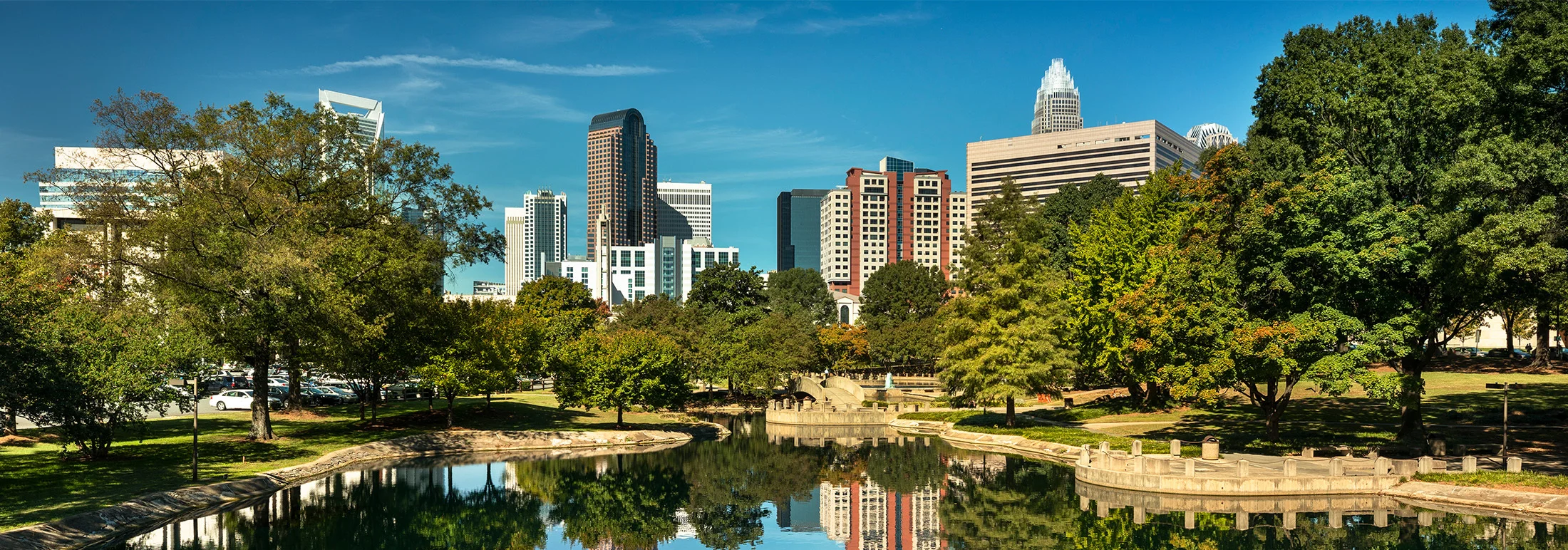 Charlotte skyline viewed from Marshall Park, featuring modern office buildings reflected in a calm pond, framed by green trees and a clear blue sky.