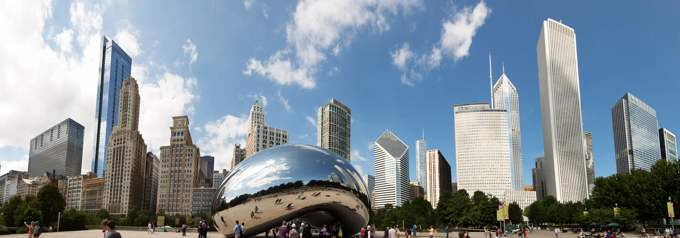 Chicago skyline with Cloud Gate sculpture (the Bean) in Millennium Park, surrounded by visitors, tall skyscrapers, trees, and a bright blue sky with scattered clouds.